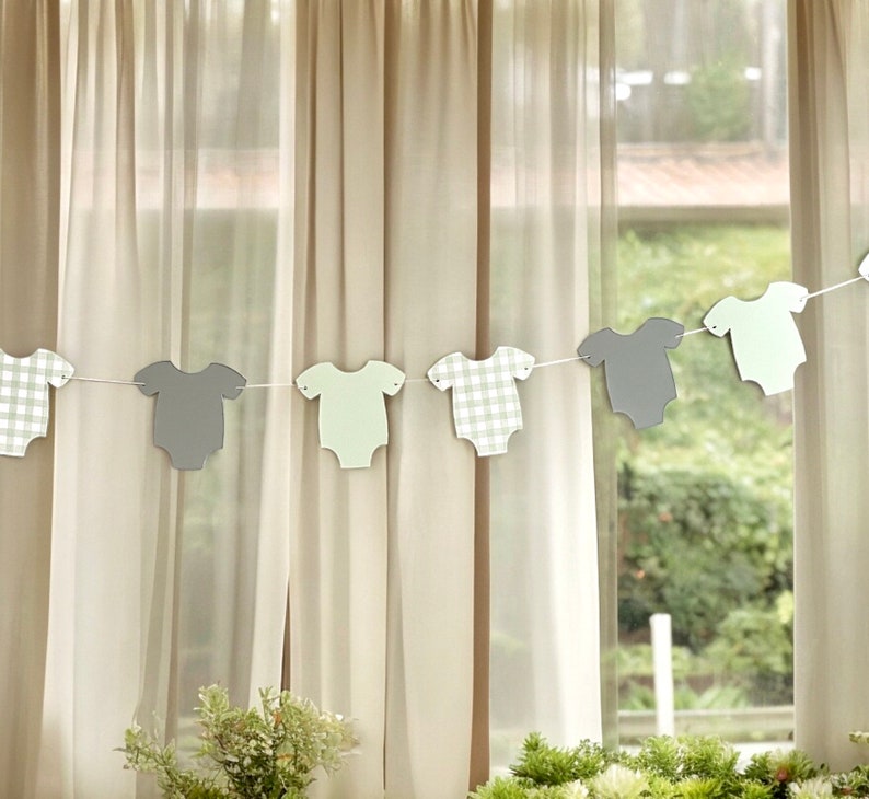 Op de afbeelding: Een baby shower banner met afwisselend grijze en groene baby rompers. De rompers zijn uitgesneden en aan elkaar geregen met wit touw.