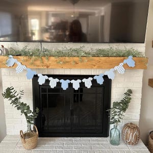 Op de afbeelding: Een blauwe en witte geruite baby romper banner hangt boven een open haard met een zwarte schouw. De banner is gemaakt van papier en heeft een string van blauwe en witte geruite baby rompers eraan bevestigd. De open haard is wit en heeft een zwart open haard scherm.