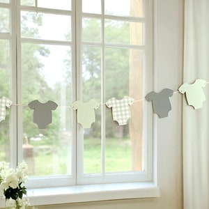 Op de afbeelding: Een baby shower banner met een string van grijze en groene papieren uitsnijdingen van rompers. De rompers hebben verschillende patronen, waaronder ruitjes en effen kleuren.