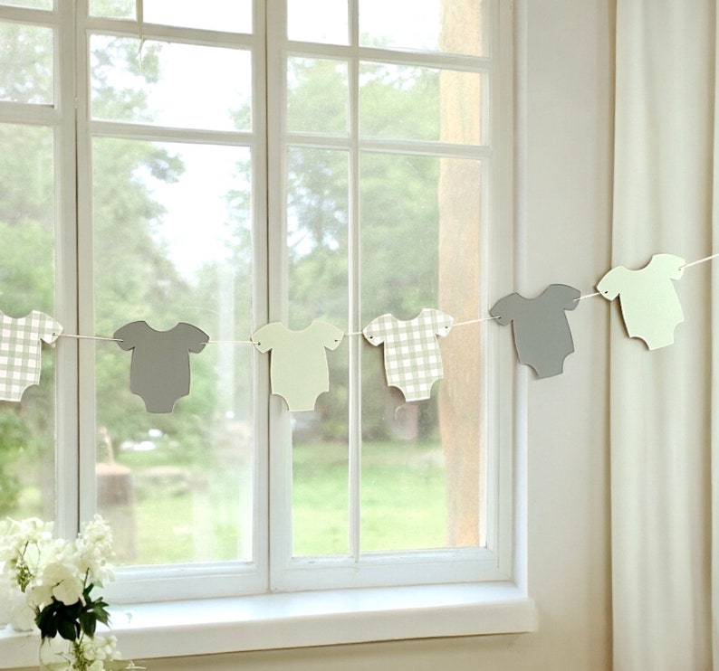 Op de afbeelding: Een baby shower banner met een string van grijze en groene papieren uitsnijdingen van rompers. De rompers hebben verschillende patronen, waaronder ruitjes en effen kleuren.