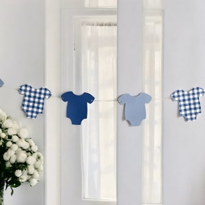 May include: A blue and white gingham baby onesie banner with alternating solid blue onesies. The banner is strung with white twine and is hanging in front of a window.