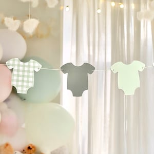 Op de afbeelding: Een baby shower banner met drie papieren uitsnijdingen van rompers in groen, grijs en wit ruitjespatroon. De rompers zijn aan elkaar geregen met wit touw.