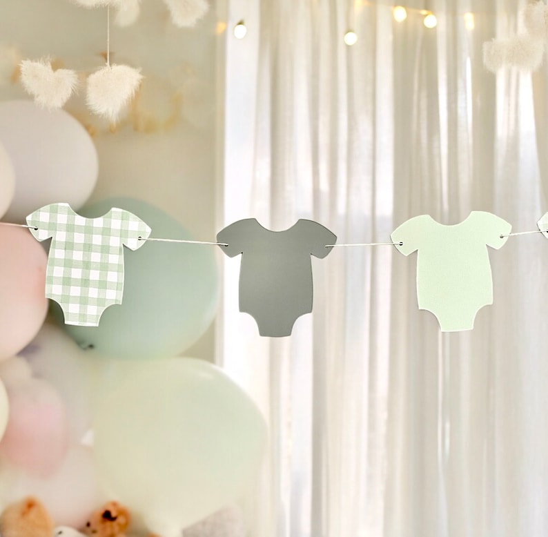Op de afbeelding: Een baby shower banner met drie papieren uitsnijdingen van rompers in groen, grijs en wit ruitjespatroon. De rompers zijn aan elkaar geregen met wit touw.