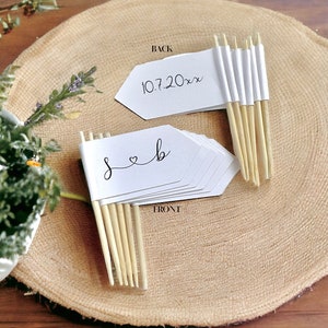 May include: White paper flags with wooden sticks, featuring the initials "S & B" with a heart and the date "10.7.20xx". The flags are arranged on a rustic wooden slice, suitable for wedding or event decor.