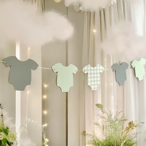 Op de afbeelding: Een baby shower banner met vijf papieren uitsnijdingen van rompertjes in grijs, groen en een groen-wit geruit patroon. De rompertjes zijn aan elkaar geregen met wit touw.