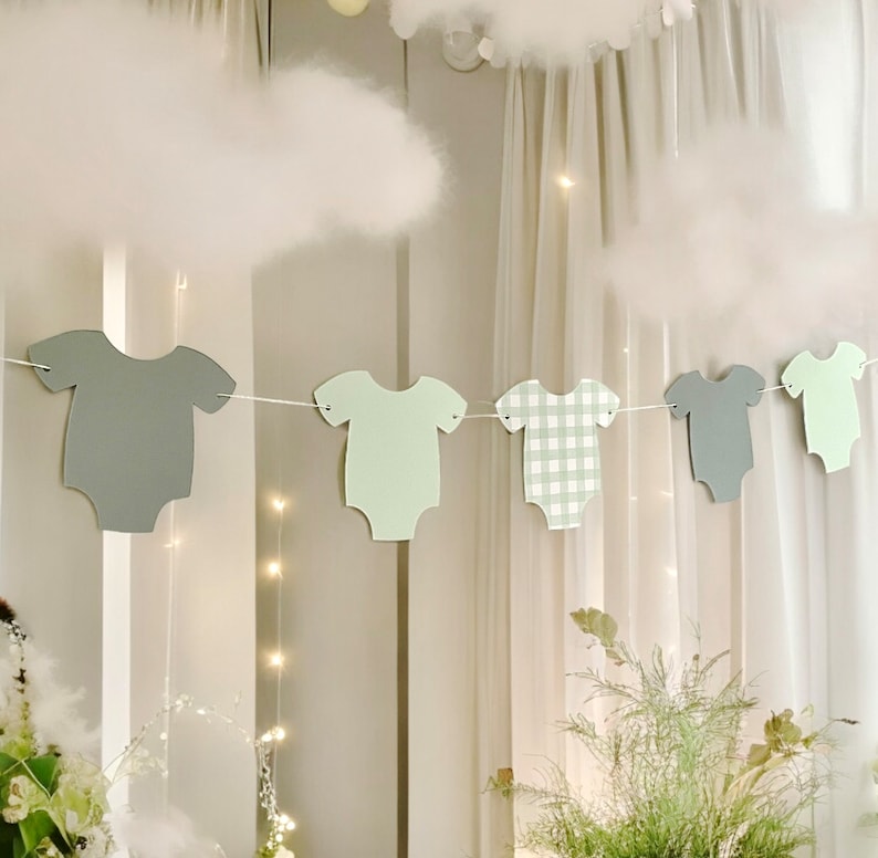 Op de afbeelding: Een baby shower banner met vijf papieren uitsnijdingen van rompertjes in grijs, groen en een groen-wit geruit patroon. De rompertjes zijn aan elkaar geregen met wit touw.