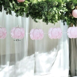 May include: A pink pumpkin garland with glitter detailing is strung across the image. The garland is made of paper and has a string attached to each pumpkin. The pumpkins are decorated with glitter and have a stem at the top. The text "-1.5-" is printed on the garland.