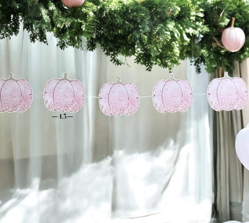 May include: A pink pumpkin garland with glitter detailing is strung across the image. The garland is made of paper and has a string attached to each pumpkin. The pumpkins are decorated with glitter and have a stem at the top. The text "-1.5-" is printed on the garland.