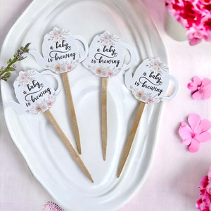 May include: Four white cupcake toppers shaped like teapots with floral designs and the text "a baby is brewing".