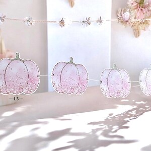 May include: A decorative garland featuring pink pumpkin shapes with glitter detailing, strung on a white cord. Each pumpkin is about 11.4 cm wide. The background is a soft, blurred white.