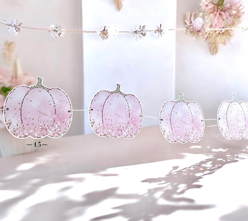 May include: A decorative garland featuring pink pumpkin shapes with glitter detailing, strung on a white cord. Each pumpkin is about 11.4 cm wide. The background is a soft, blurred white.