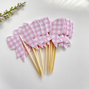May include: A collection of pink and white gingham bow cupcake toppers. Each bow is attached to a wooden skewer. The toppers are arranged on a white surface, with a sprig of lavender in the background. These are ideal for a baby shower or birthday party.