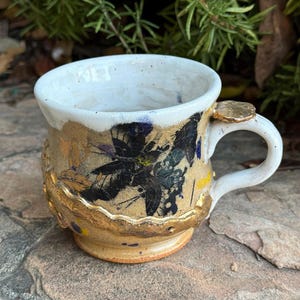 Puede incluir: Una taza de cerámica con interior blanco y exterior texturizado de color beige, con patrones florales negros y detalles dorados. La taza tiene un asa curva con un pequeño detalle circular plano de color dorado en el punto de unión superior.