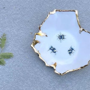 Puede incluir: Un cuenco de cerámica blanca con forma orgánica irregular y un borde pintado en oro metálico. El interior del plato presenta tres pequeñas ilustraciones detalladas de abejorros dispuestas en un patrón triangular sobre la superficie blanca.