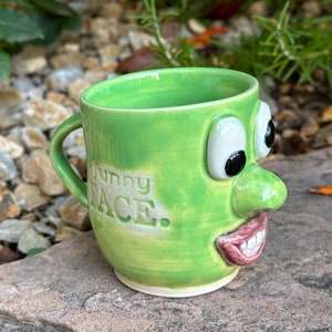 Op de afbeelding: Een groene keramische mok met een grappig gezicht en de woorden "Funny Face". De mok heeft een grote neus, grote ogen en een brede glimlach.