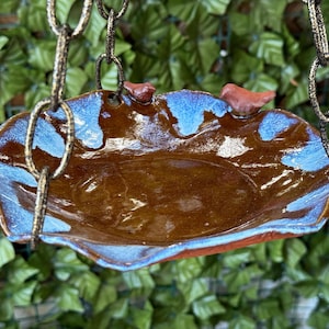 Puede incluir: Un bebedero para pájaros de cerámica marrón con un esmalte azul y una cadena para colgar. El bebedero tiene un borde festoneado y está decorado con pequeños pájaros rojos.