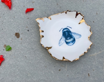 May include: A small white ceramic dish with a gold rim and a blue and white bee illustration. The dish is on a gray surface.
