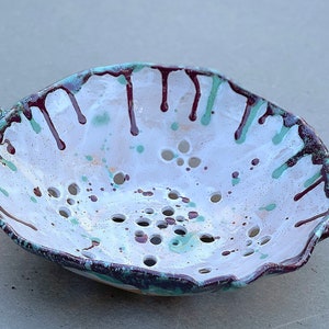 May include: White ceramic colander with two handles, decorated with brown and green paint splatters. The colander has a scalloped edge and a pattern of small holes.
