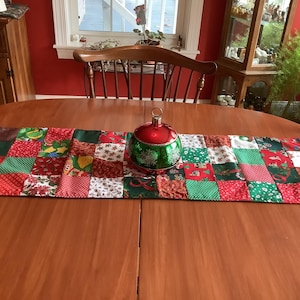May include: A festive Christmas table runner with a patchwork design in red, green, and white fabrics. A large, round, red and green ornament sits atop the runner. The runner is trimmed with red pom-poms.