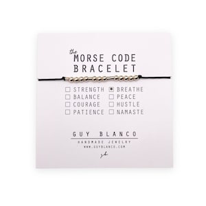 May include: A black string bracelet with silver beads and a silver bar in the center. The bracelet is displayed on a white card with the text "the MORSE CODE BRACELET" and a list of words that the beads represent: "STRENGTH", "BREATHE", "BALANCE", "PEACE", "COURAGE", "HUSTLE", "PATIENCE", "NAMASTE". The card also includes the text "GUY BLANCO" and "HANDMADE JEWELRY WWW.GUYBLANCO.COM".