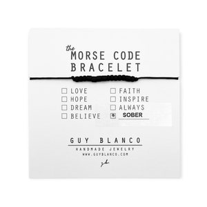 May include: A black string bracelet with black beads. The bracelet is displayed on a white card with the words "the MORSE CODE BRACELET" and a list of words that can be spelled out using the beads, including "LOVE", "HOPE", "DREAM", "BELIEVE", "FAITH", "INSPIRE", "ALWAYS", and "SOBER". The card also includes the text "GUY BLANCO HANDMADE JEWELRY WWW.GUYBLANCO.COM".
