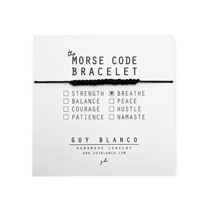 May include: Black string bracelet with black beads. The bracelet is on a white card with the text "the MORSE CODE BRACELET" and a list of words: STRENGTH, BALANCE, COURAGE, PATIENCE, BREATHE, PEACE, HUSTLE, NAMASTE. The card also includes the text "GUY BLANCO" and "HANDMADE JEWELRY WWW.GUYBLANCO.COM".