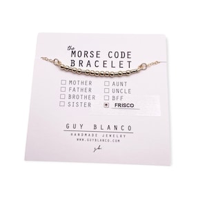 May include: A gold Morse code bracelet on a white card with the text "the MORSE CODE BRACELET" and a list of options for who the bracelet is for, including "Mother", "Father", "Brother", "Sister", "Aunt", "Uncle", "BFF", and "Frisco". The card also includes the text "GUY BLANCO HANDMADE JEWELRY WWW.GUYBLANCO.COM".