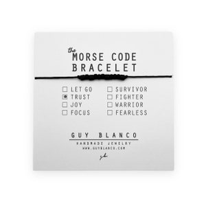Puede incluir: Pulsera de cuerda negra con cuentas negras. La pulsera se muestra en una tarjeta blanca con el texto "the MORSE CODE BRACELET" y una lista de palabras que se pueden deletrear usando las cuentas. Las palabras son "LET GO", "TRUST", "JOY", "FOCUS", "SURVIVOR", "FIGHTER", "WARRIOR" y "FEARLESS". La tarjeta también incluye el texto "GUY BLANCO HANDMADE JEWELRY WWW.GUYBLANCO.COM".