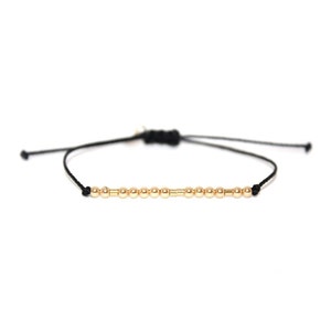 May include: A simple gold bead bracelet with a black cord. The bracelet is adjustable and can be worn on the wrist or ankle.