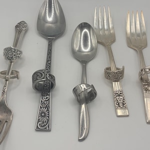 May include: Silverware set featuring spoons and forks, each adorned with decorative rings. The spoons and forks have intricate designs, including floral and geometric patterns. The silverware is arranged on a white surface.