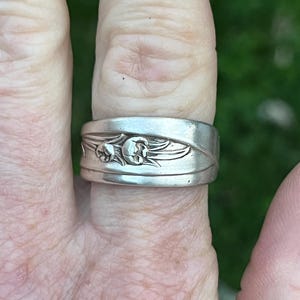 May include: A silver spoon ring with a floral design. The ring is worn on a finger.