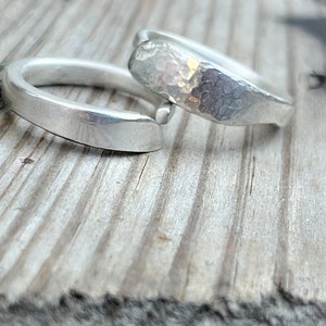 May include: Two silver rings, one with a smooth finish and the other with a hammered texture, are stacked on top of each other on a wooden surface.