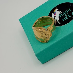 May include: A gold-colored spoon ring with a floral design. The ring is set on a teal-colored box with a black sticker that reads "MADE BY MELZ" and features a unicorn.