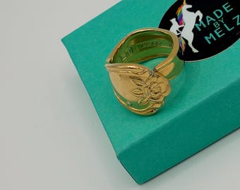 May include: A gold-colored spoon ring with a floral design. The ring is set on a teal-colored box with a black sticker that reads "MADE BY MELZ" and features a unicorn.