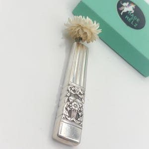 May include: A silver-colored, rectangular vase with a floral design and a dried flower. The vase has vertical lines and a rectangular base. A light green box with a unicorn sticker is in the background.