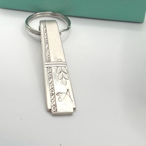 May include: Silver-toned metal keychain with a rectangular pendant. The pendant features engraved floral and leaf designs, with the letter "A" etched on the bottom. A silver key ring is attached to the top of the pendant. The background includes a teal box.