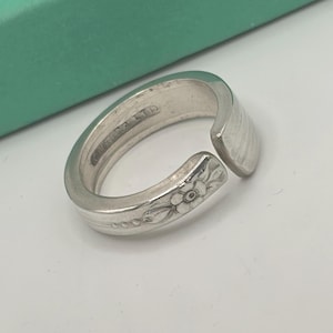 May include: A silver ring crafted from a spoon, featuring a floral design and the inscription "ONEIDA LTD." The ring has a curved shape with an open design, showcasing the repurposed silverware aesthetic. The ring is on a white background.