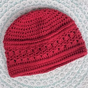 Puede incluir: Un gorro de ganchillo rojo hecho a mano. El gorro presenta un patr&oacute;n de puntadas texturizadas con una banda decorativa alrededor del centro. El gorro est&aacute; hecho de un hilo suave y c&aacute;lido, ideal para el fr&iacute;o. El gorro se muestra sobre una superficie blanca y de encaje.