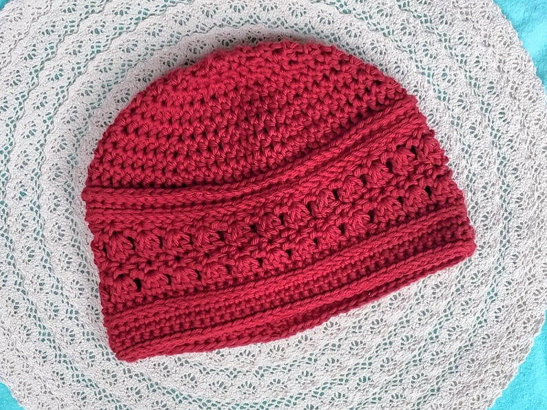 Puede incluir: Un gorro de ganchillo rojo hecho a mano. El gorro presenta un patr&oacute;n de puntadas texturizadas con una banda decorativa alrededor del centro. El gorro est&aacute; hecho de un hilo suave y c&aacute;lido, ideal para el fr&iacute;o. El gorro se muestra sobre una superficie blanca y de encaje.