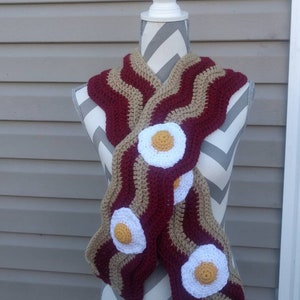 Puede incluir: Una bufanda de ganchillo con un patr&oacute;n ondulado en rayas alternas de color granate oscuro y beige, dise&ntilde;ada para parecerse a tiras de tocino. La bufanda est&aacute; decorada con tres aplicaciones circulares de ganchillo en blanco y amarillo.