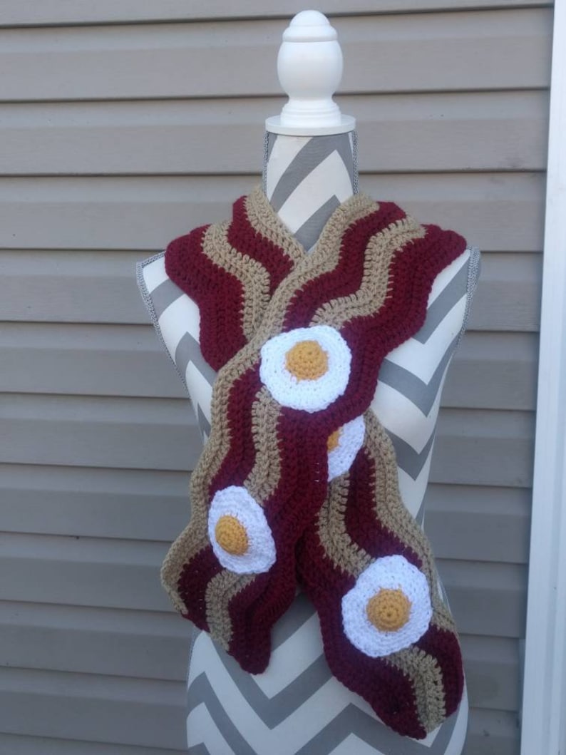 Puede incluir: Una bufanda de ganchillo con un patr&oacute;n ondulado en rayas alternas de color granate oscuro y beige, dise&ntilde;ada para parecerse a tiras de tocino. La bufanda est&aacute; decorada con tres aplicaciones circulares de ganchillo en blanco y amarillo.