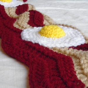Puede incluir: Una bufanda de ganchillo con un patr&oacute;n repetitivo de tiras de tocino rojo oscuro y capas de grasa beige, acentuada con motivos tridimensionales de huevo frito que consisten en claras blancas y yemas amarillas en relieve cosidas.