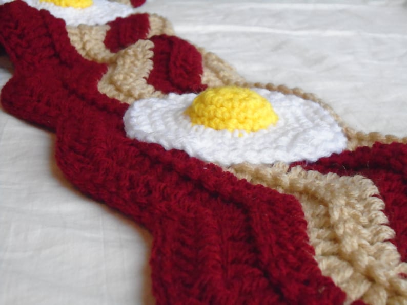 Puede incluir: Una bufanda de ganchillo con un patr&oacute;n repetitivo de tiras de tocino rojo oscuro y capas de grasa beige, acentuada con motivos tridimensionales de huevo frito que consisten en claras blancas y yemas amarillas en relieve cosidas.