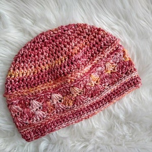 May include: A hand-crocheted beanie hat featuring a variegated yarn pattern in shades of deep red, magenta, and soft orange. The design incorporates a textured stitch pattern around the lower section, transitioning into a simple stitch at the top.