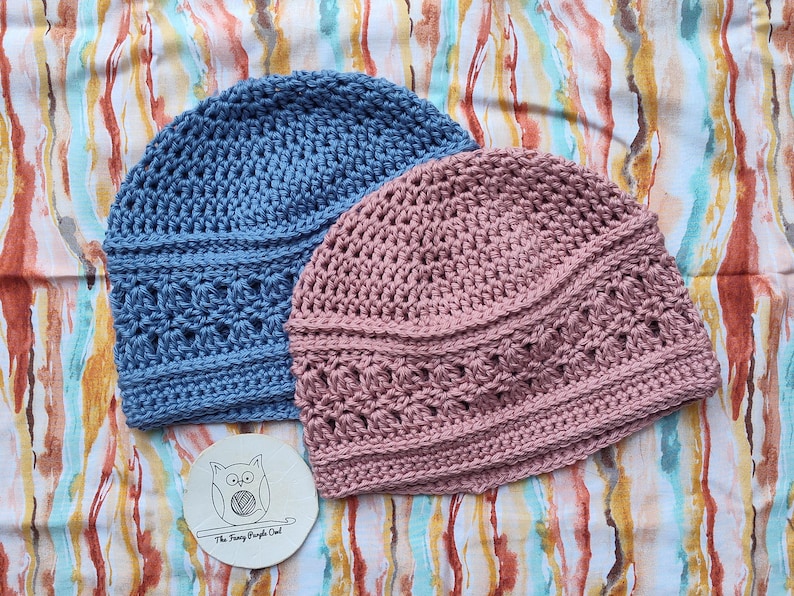 Puede incluir: Dos gorros de ganchillo hechos a mano en azul y rosa empolvado. Los gorros tienen un patr&oacute;n de puntada texturizada y se muestran sobre una tela estampada colorida. Tambi&eacute;n hay un peque&ntilde;o posavasos redondo con una ilustraci&oacute;n de b&uacute;ho y el texto "The Funky Purple Owl".