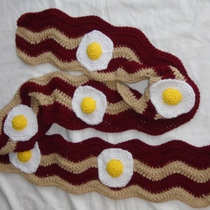 Puede incluir: Una bufanda larga de ganchillo con un patr&oacute;n ondulado de rayas en granate oscuro y beige, que recuerda a tiras de tocino. La bufanda est&aacute; decorada con cinco aplicaciones circulares de ganchillo en blanco y amarillo con forma de huevo.