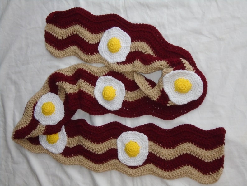 Puede incluir: Una bufanda larga de ganchillo con un patr&oacute;n ondulado de rayas en granate oscuro y beige, que recuerda a tiras de tocino. La bufanda est&aacute; decorada con cinco aplicaciones circulares de ganchillo en blanco y amarillo con forma de huevo.