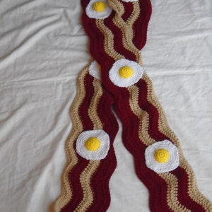Puede incluir: Una bufanda larga de ganchillo con un patr&oacute;n ondulado de rayas en rojo oscuro y beige, que imita la apariencia de tiras de tocino. La bufanda est&aacute; decorada con varios apliques circulares de ganchillo blancos y amarillos como huevos fritos.