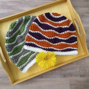 May include: Two crocheted hats in a wooden tray. One hat is green, white, and gray with a wavy pattern. The other hat is orange, white, and purple with a wavy pattern. A yellow flower is in the tray.