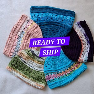 May include: Five crocheted beanies arranged in a circle, featuring various colors including teal, pink, olive green, bright blue, and dark purple. Each hat displays a textured stitch pattern and a decorative band. The text READY TO SHIP is centered.